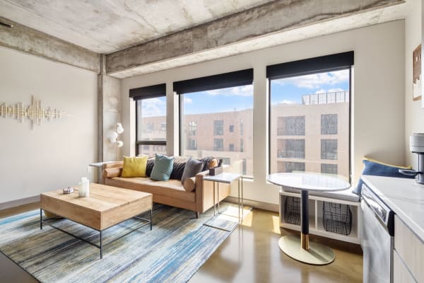 Modern living room with a leather sofa, colorful cushions, and large windows overlooking an urban view.
