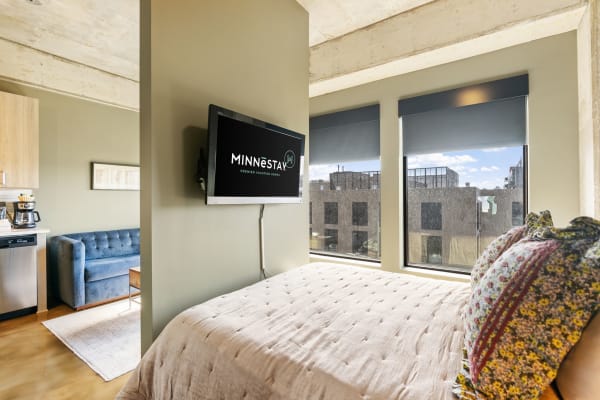 A stylish bedroom featuring a bed, a TV with MINNESTAY logo, and a cozy blue couch.