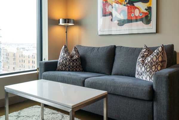 Modern living room with a gray sofa and decorative pillows, window view of city, and a lamp.