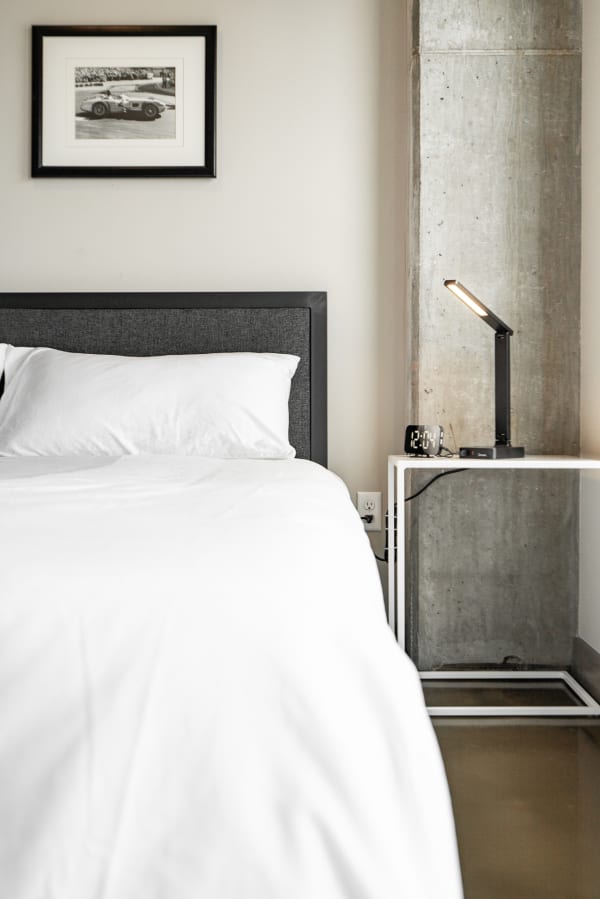 A modern bedroom featuring a gray headboard, white linens, and a minimalist bedside table with lamp and clock.