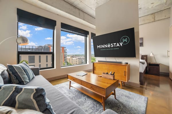 Modern living room with a sofa, city views from windows, and a TV displaying MINNēSTAY logo.