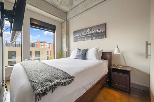 A cozy modern bedroom featuring a bed with white linens, a decorative throw, large windows, and an urban skyline picture.