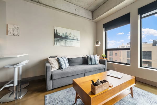 A modern living room featuring a gray sofa, art on the walls, and large windows.