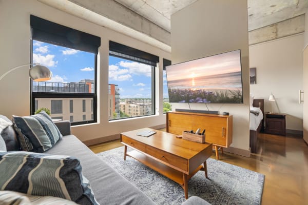Modern living room with a gray sofa, wooden coffee table, large windows, and a view of blue skies.