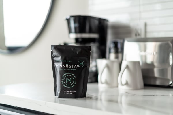 Black coffee bag labeled MINNESTAY on a marble countertop with a coffee maker and white mugs in the background.