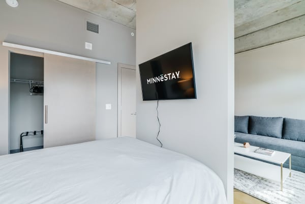 Modern bedroom with a bed, a wall-mounted TV displaying 'MINNēSTAY', and a gray sofa in the background.