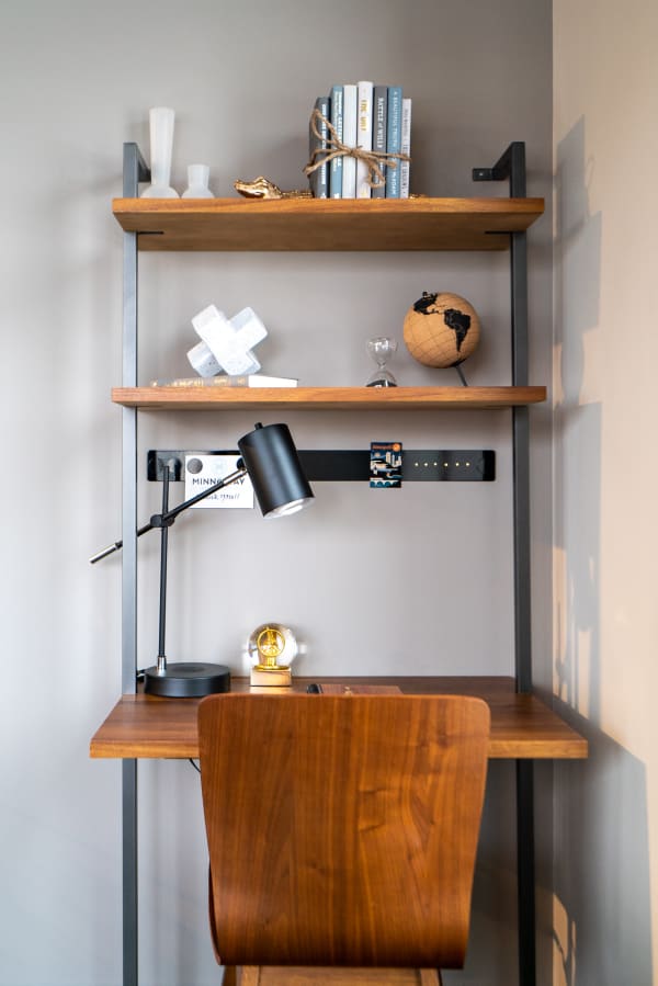 A home office with a wooden desk, shelves containing books and decor, and a lamp.