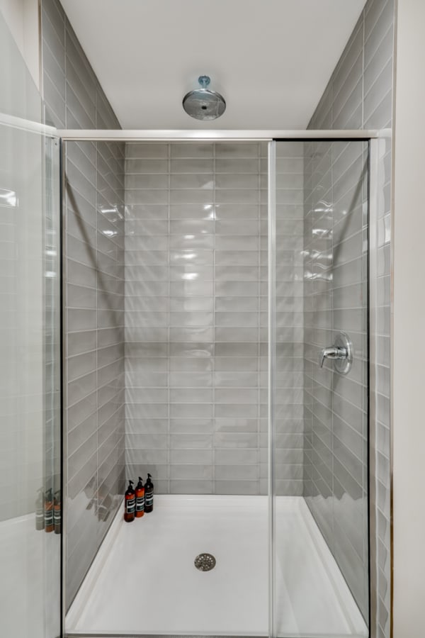 A clean shower space with gray tiles and a glass door, featuring toiletries on the floor.