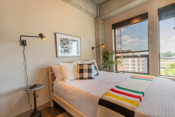 A stylish bedroom with a bed, decorative cushions, a side table, and large windows overlooking an urban landscape.