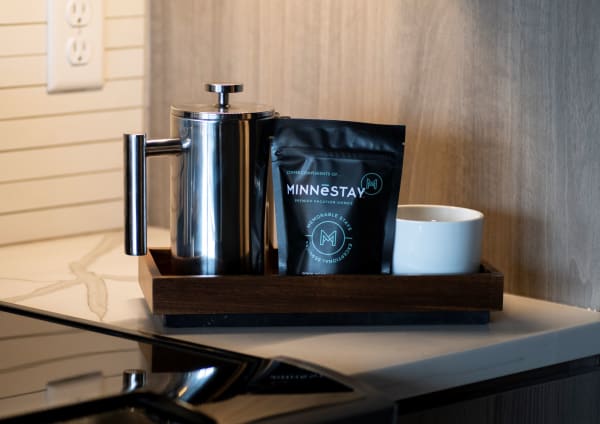 A stainless steel French press, black coffee bag labeled MINNESTAY, and a white cup on a wooden tray on a marble countertop.