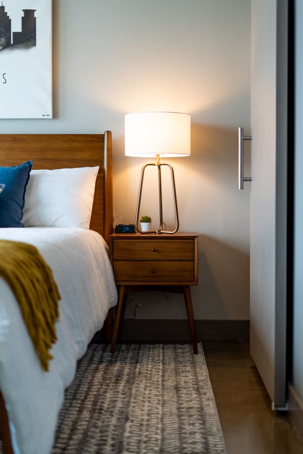 A cozy bedroom corner showing a wooden bed, nightstand with lamp and clock, and patterned rug.