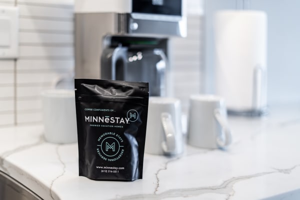 Minnēstay coffee bag on a marble countertop with white ceramic mugs in soft focus.