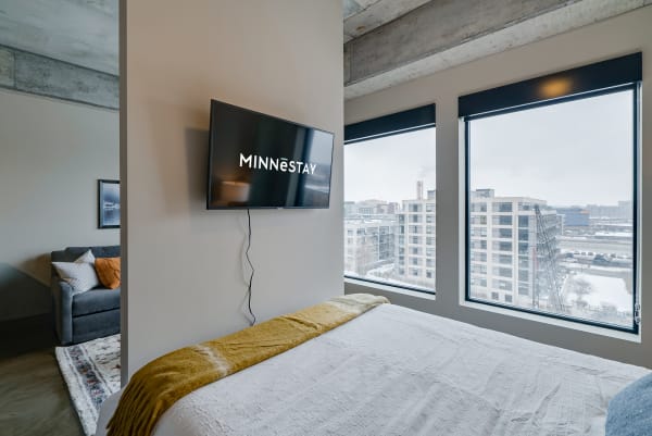 Modern bedroom featuring a bed with white bedding, a mustard throw, and a television displaying 'MINNēSTAY', with large windows offering a snowy city landscape.