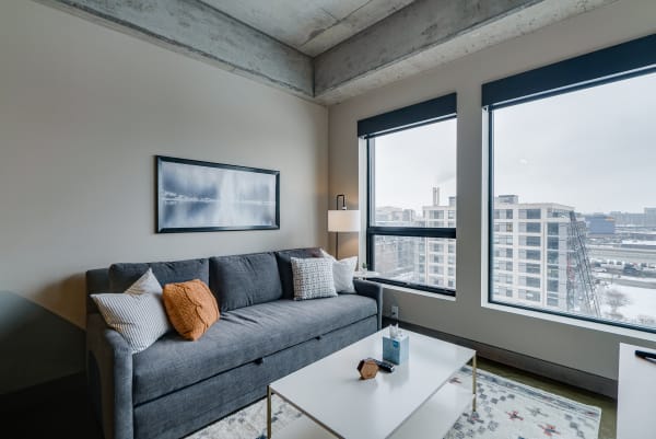 Contemporary living room with gray sofa and urban view through large windows.