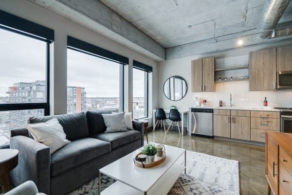 A contemporary living space with a gray sofa, coffee table, and kitchen area, featuring large windows and a city view.