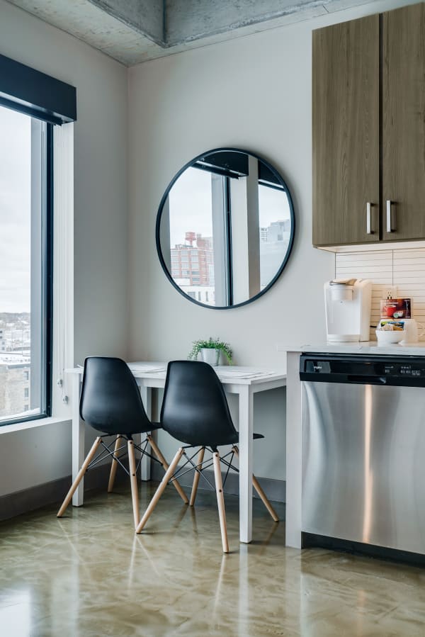 A modern dining area with a white table, two black chairs, a round mirror, and a view of the city through large windows.