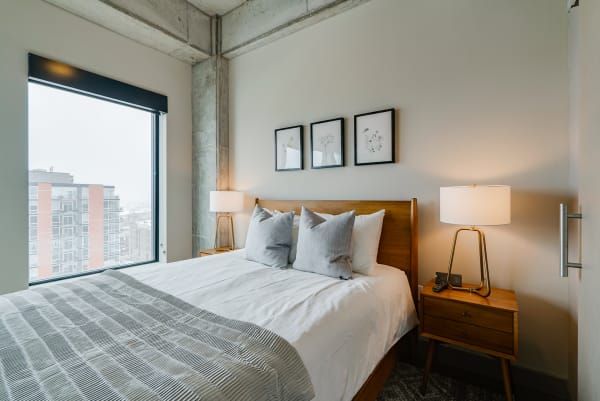 A cozy bedroom with a large window, soft bedding, and botanical prints on the wall.