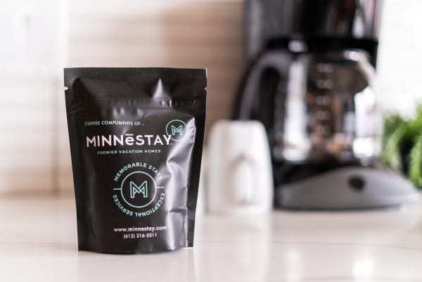 A coffee package with the MINNESTAY logo in front of a coffee maker and a sugar bowl.