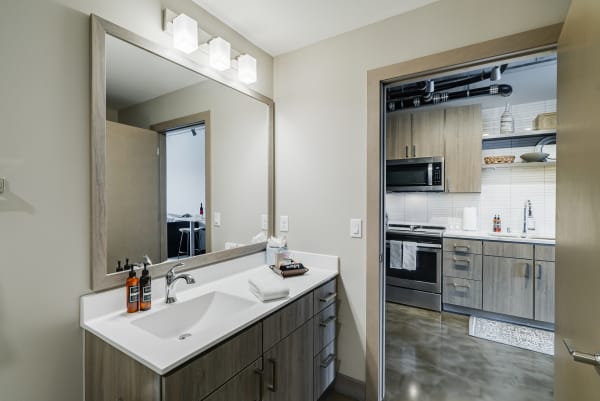 A contemporary bathroom featuring a white sink, wooden cabinets, and a view into a kitchen with gray cabinets.