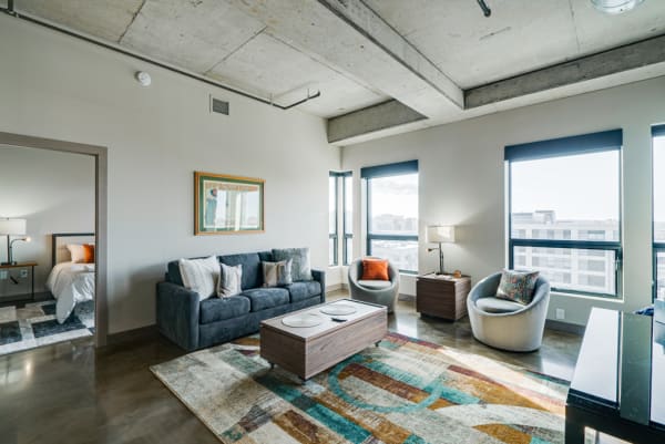 A stylish living room featuring a grey sofa, colorful rugs, and large windows.