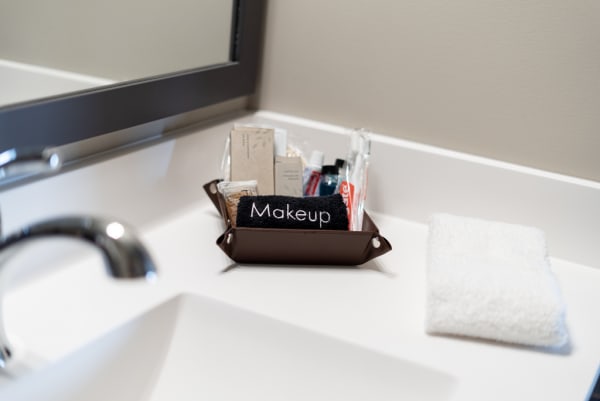 A brown tray with makeup products labeled 'Makeup' next to a folded white washcloth on a bathroom counter.