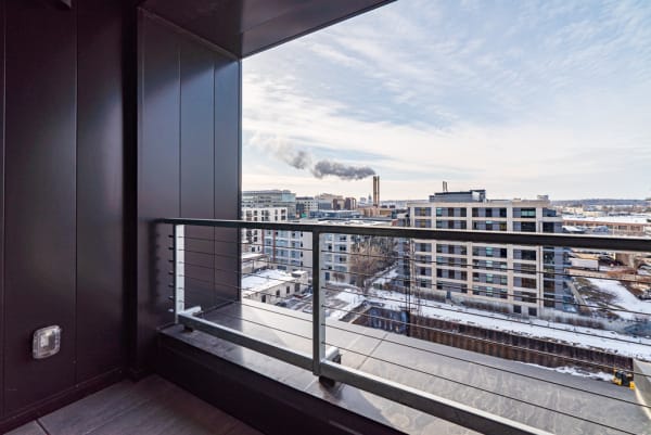 A modern balcony with a view of snowy urban buildings and a smokestack emitting smoke against a cloudy sky.