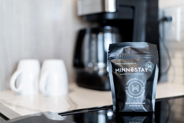 Black coffee package with MINNESTAY branding next to a coffee maker and white mugs on a kitchen counter.