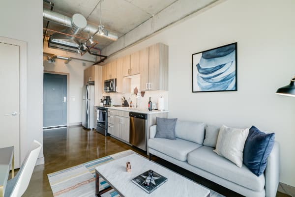 A modern apartment with a kitchen and living area featuring light wooden cabinets, a gray sofa, and vibrant artwork.