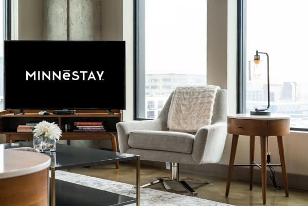 Contemporary living room with gray armchair, side table, and television showing MINNĒSTAY logo.