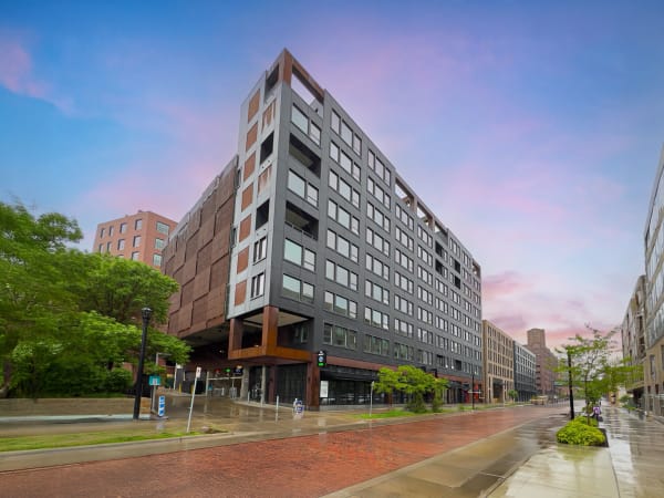 A contemporary building with dark gray and rust-colored cladding in an urban setting, with a brick-paved street and greenery.