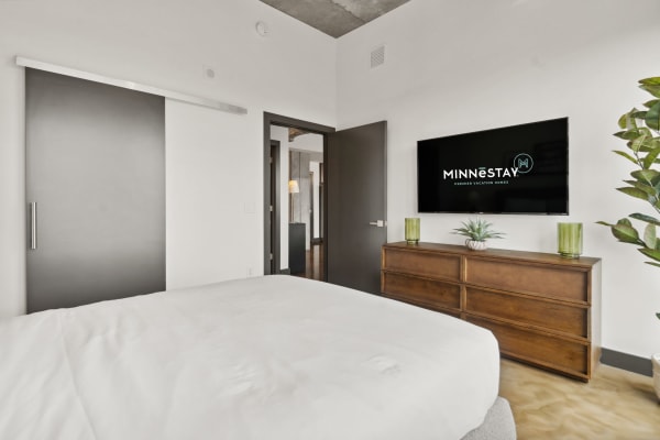 Modern bedroom with a bed, TV displaying MINNESTAY logo, and a wooden dresser with plants.