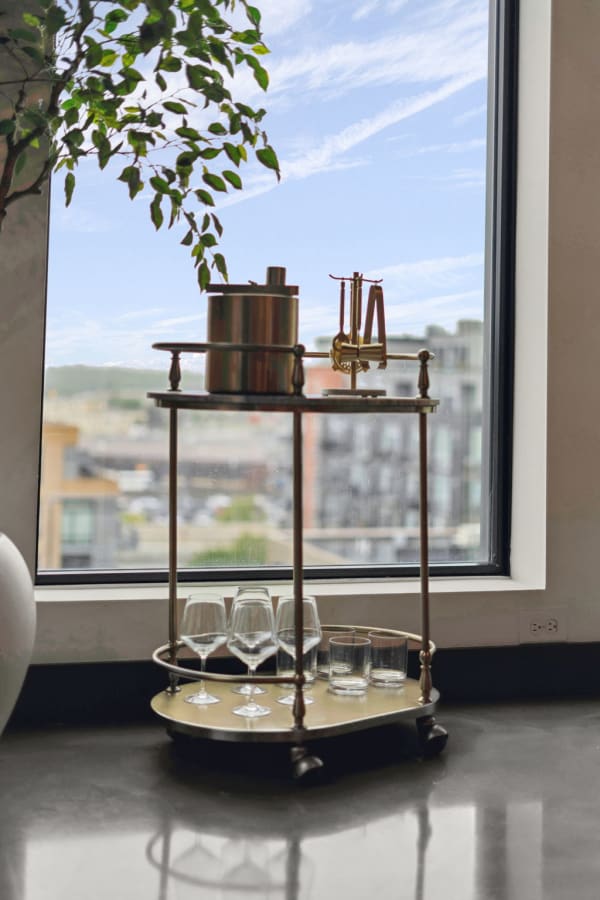 A bar cart with glassware and an ice bucket is positioned next to a large window showing a city view.