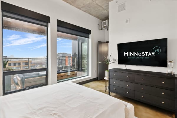 Modern bedroom with large windows and MINNĒSTAY branding on the TV.