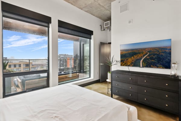 Modern bedroom featuring large windows, a black dresser with TV, and a soft bed.