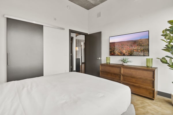 Modern bedroom with a white bed, wooden dresser, and wall-mounted TV showing an autumn landscape.