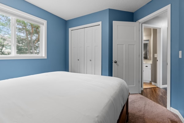 Cozy bedroom with blue walls and white bedding, featuring a window and adjoining bathroom entrance.