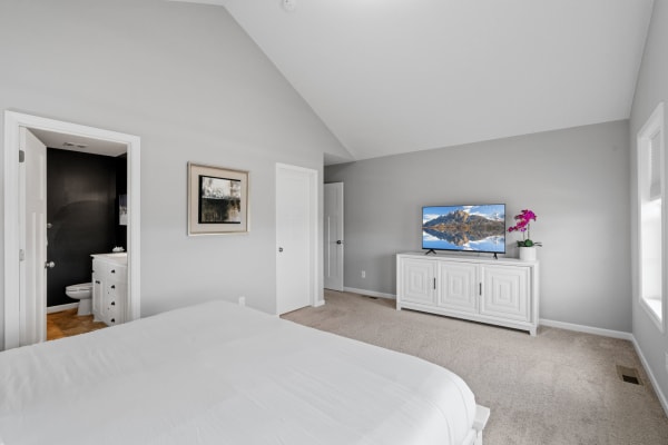 Spacious bedroom with a high ceiling, white bedspread, and a television displaying a landscape.