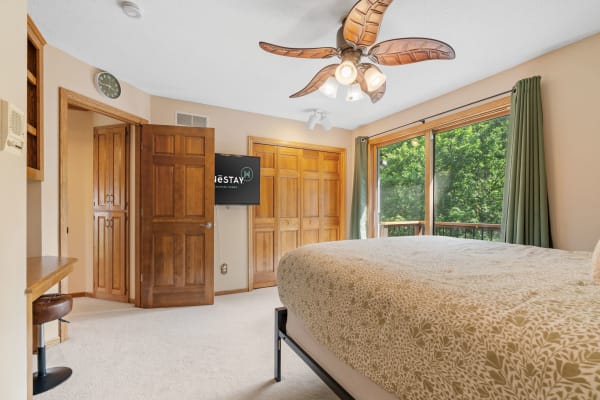 A modern bedroom with a large bed, ceiling fan, television, and sliding glass door to a green view.