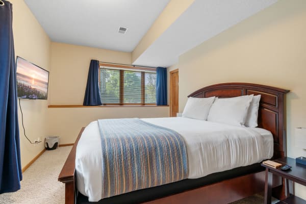 A cozy bedroom featuring a bed with a striped blanket, a mounted TV, and a window with blue curtains.