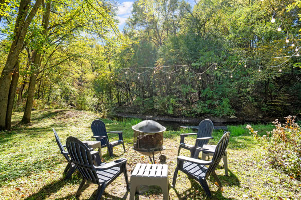 Outdoor seating area with a fire pit and Adirondack chairs in a sunny forest environment.