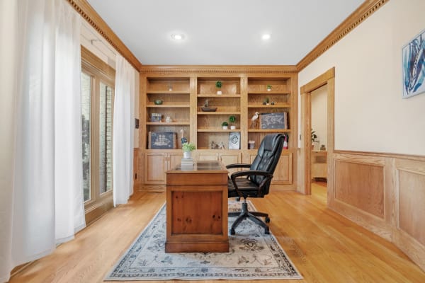 Office interior featuring a wooden desk, black chair, built-in shelves, and natural light through sheer curtains.