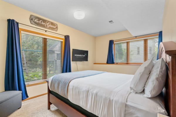 A comfortable bedroom featuring a neatly made bed, dark blue curtains, a mounted TV, and a decorative wall sign.