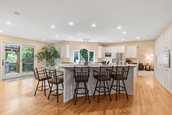 An open kitchen with an island, bar stools, white cabinets, and large windows overlooking a wooded area.