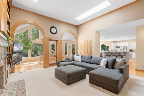 Living area featuring a gray sectional couch, TV, and large windows with a dining area and kitchen in view.