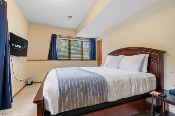 Cozy bedroom featuring a large bed, a mounted television displaying Minnestay, and a window with navy curtains.