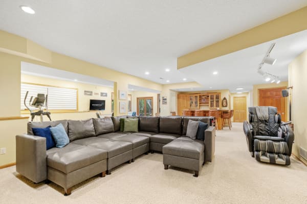 Spacious basement featuring a grey sectional couch, exercise equipment, and a bar area in the background.