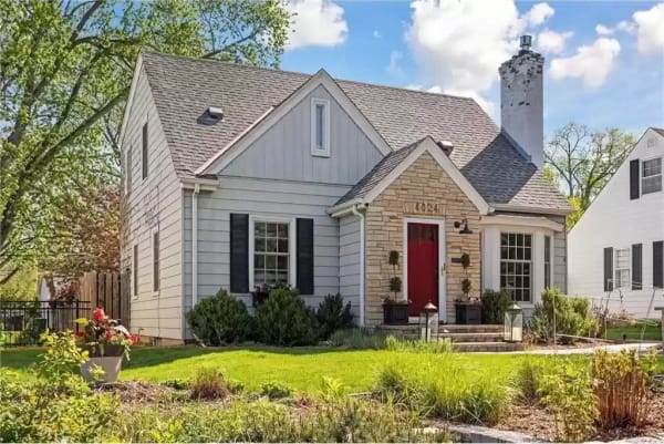 Exterior view of a charming house with a stone façade and red door, surrounded by greenery.