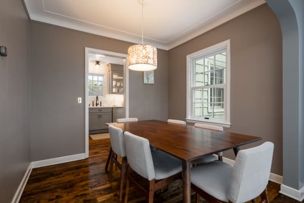 Modern dining room with wooden table and gray upholstered chairs, leading to a kitchen.