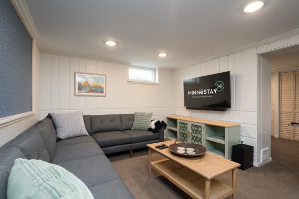 A cozy living room featuring a grey sectional couch, a light wood coffee table, and a mounted TV with MINNESTAY logo.