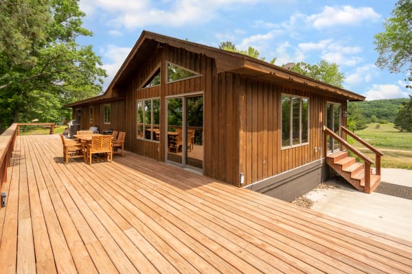 Wooden cabin with a large deck and outdoor furniture, nestled in greenery.